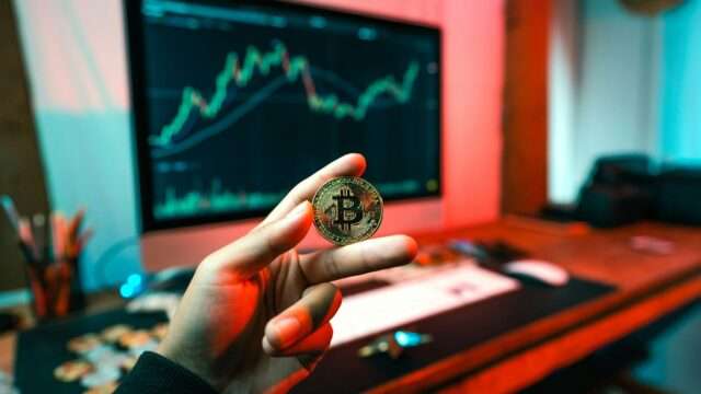 A hand holding a Bitcoin token in front of a cryptocurrency price chart