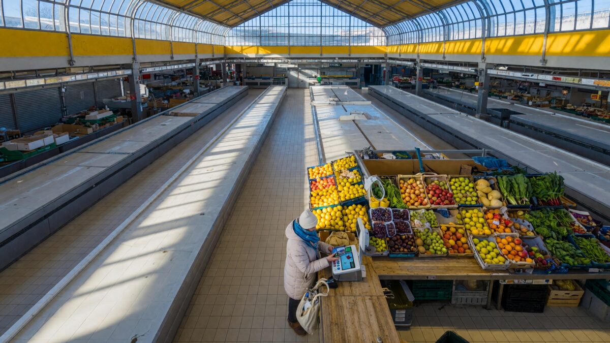 A single customer paying at a produce stall in a bright covered market hall with a contactless payment terminal on the counter