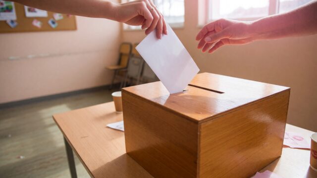 Hands dropping a folded blank paper into a polished wooden ballot box on a table in a bright community hall